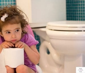 Toddler girl holding toilet paper sitting next to toilet. This article discusses the details of potty training twins in 3 days.