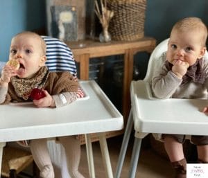 Twin babies in high chairs eating a rice cracker. This article discusses what feeding twins solids is like.