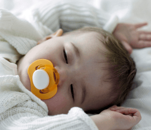 Baby with pacifier in mouth and hands above head sleeping. This article discusses how to get a 6 month old to sleep.