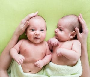Newborn twin babies laying down with an adult hand on each of their heads. This article discusses how to prepare for twins.