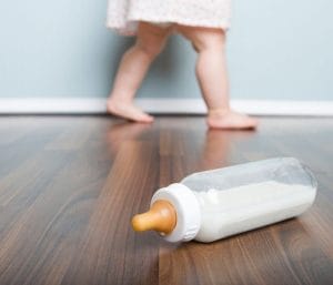 Baby bottle on wood floor with baby walking by. This section is for helping parents with their feeding baby questions.