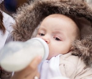 Baby in warm coat drinking bottle. This post is about how to warm bottles on the go.