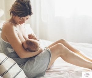 Mom nursing a baby on a bed. This post discusses when to take a breastfeeding class.