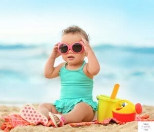 Toddler with sunglasses sitting on a towel at the beach with toys. This article discusses the things to take to the beach with a toddler.