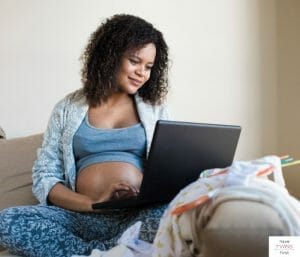Pregnant woman sitting on couch and looking at her laptop with baby clothes laying on the arm rest. This article discusses when to send out your baby registry.