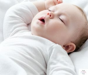 Baby with white long-sleeve onesie on and sleeping on back. This article discusses what to do when your baby is too big for a bassinet, but not ready for a crib.