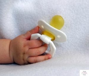 Baby's hand holding a pacifier on top of a white blanket. This article answers the questions "how many pacifiers do I need?"