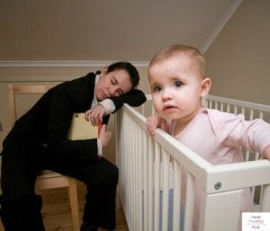 Toddler standing in crib with parent in chair asleep in background. This article discusses sleep sacks that toddler's can't get out of.