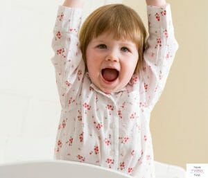 Toddler standing in crib with hands raised above head. This article discusses what to do when an overtired toddler won't sleep.