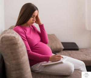 Pregnancy woman sitting on couch and writing in a notebook. This article discusses printable pregnancy checklists.
