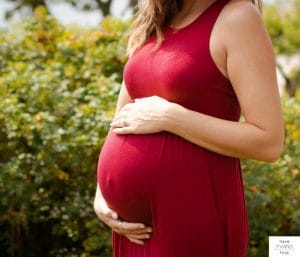 Woman wearing red dress and putting hands on pregnant belly. This article discusses what it's like at 32 weeks pregnant with twins.