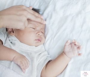 Adult checking temperature with fingers on forehead of sleeping baby. This article discusses sleep training when a baby is sick.
