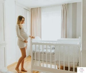 Pregnant woman standing next to a white crib in a bedroom. This article discusses a mini crib vs a regular crib.