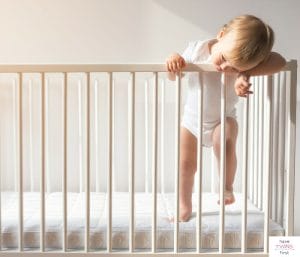Toddler standing in crib and resting head on railing. This article discusses when to lower a crib.