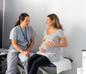 Pregnant woman sitting in exam room and and talking to a doctor. This article discusses what's on a third trimester checklist.