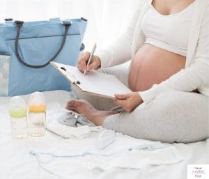 Pregnant woman holding a clipboard and pen, while sitting next to a large tote bag and baby items. This article discusses what's on a twins hospital bag checklist.