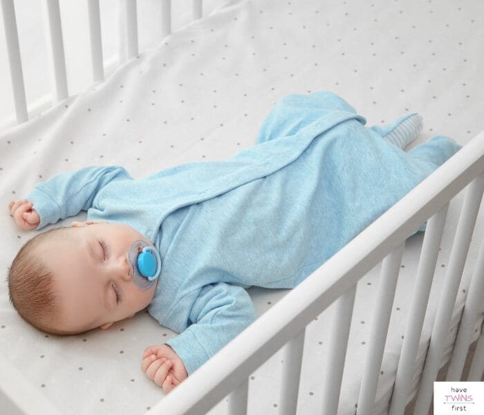 Baby wearing a blue sleeper and sleeping in a crib. This article is a baby sleeping clothes guide for a baby's first year of life.